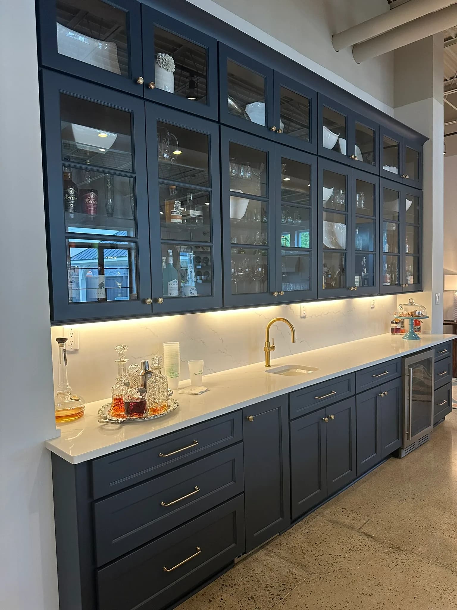 Navy bar cabinetry with glass-front uppers, a brass faucet, and a quartz counter