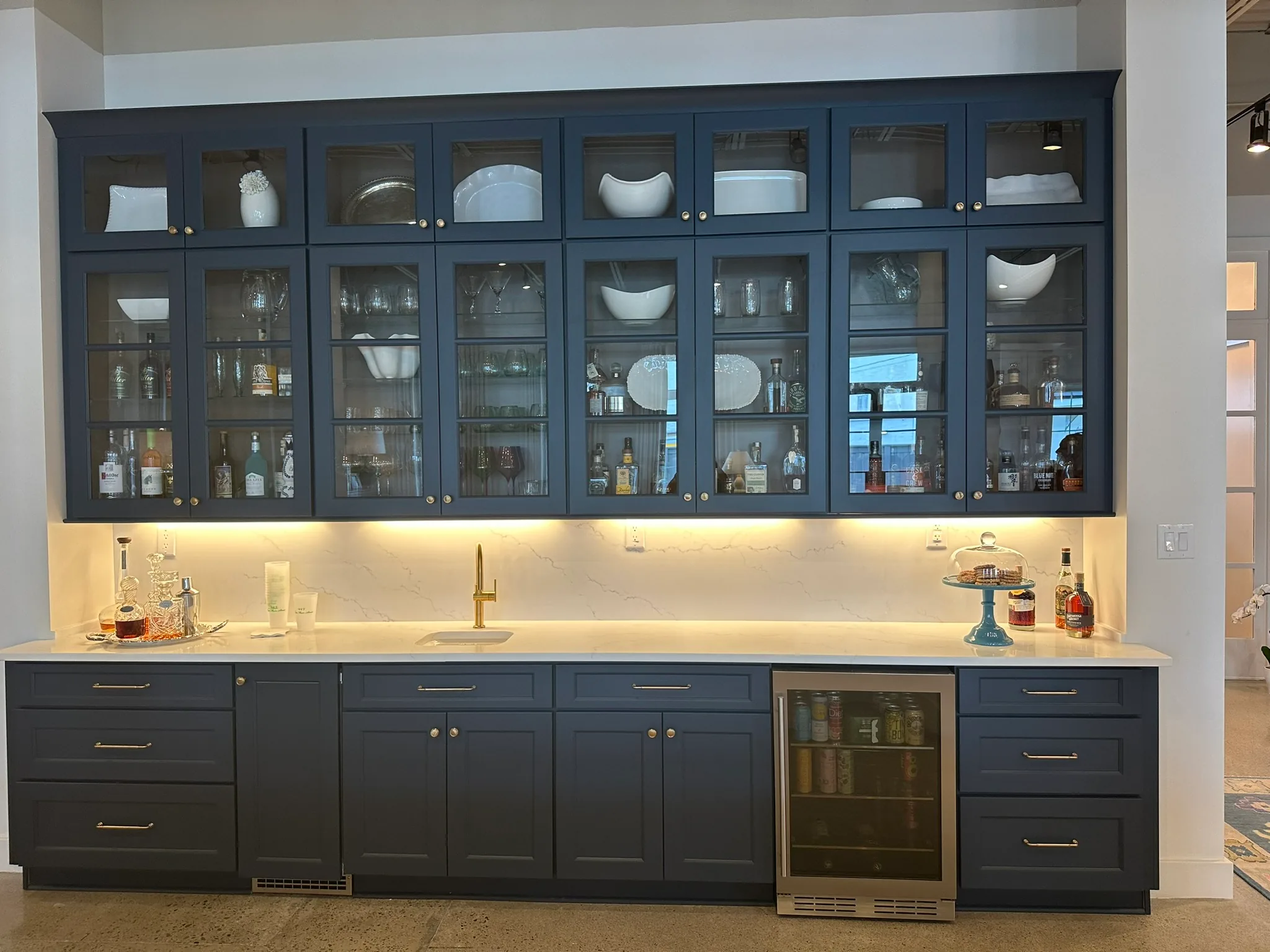 Navy wet bar with glass-front upper cabinets, under-cabinet lighting, and a beverage refrigerator