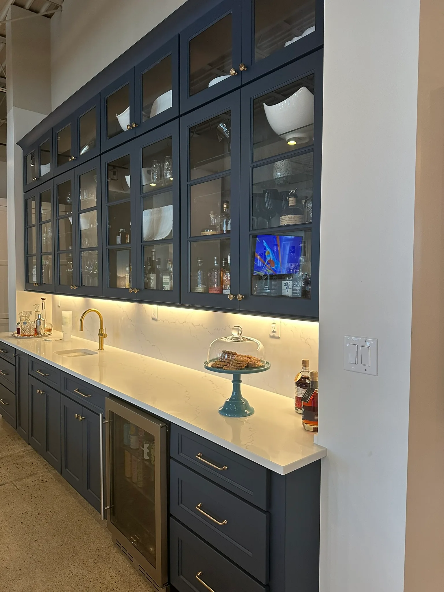 Lit glass-front bar cabinets in navy with brass hardware and a beverage refrigerator below